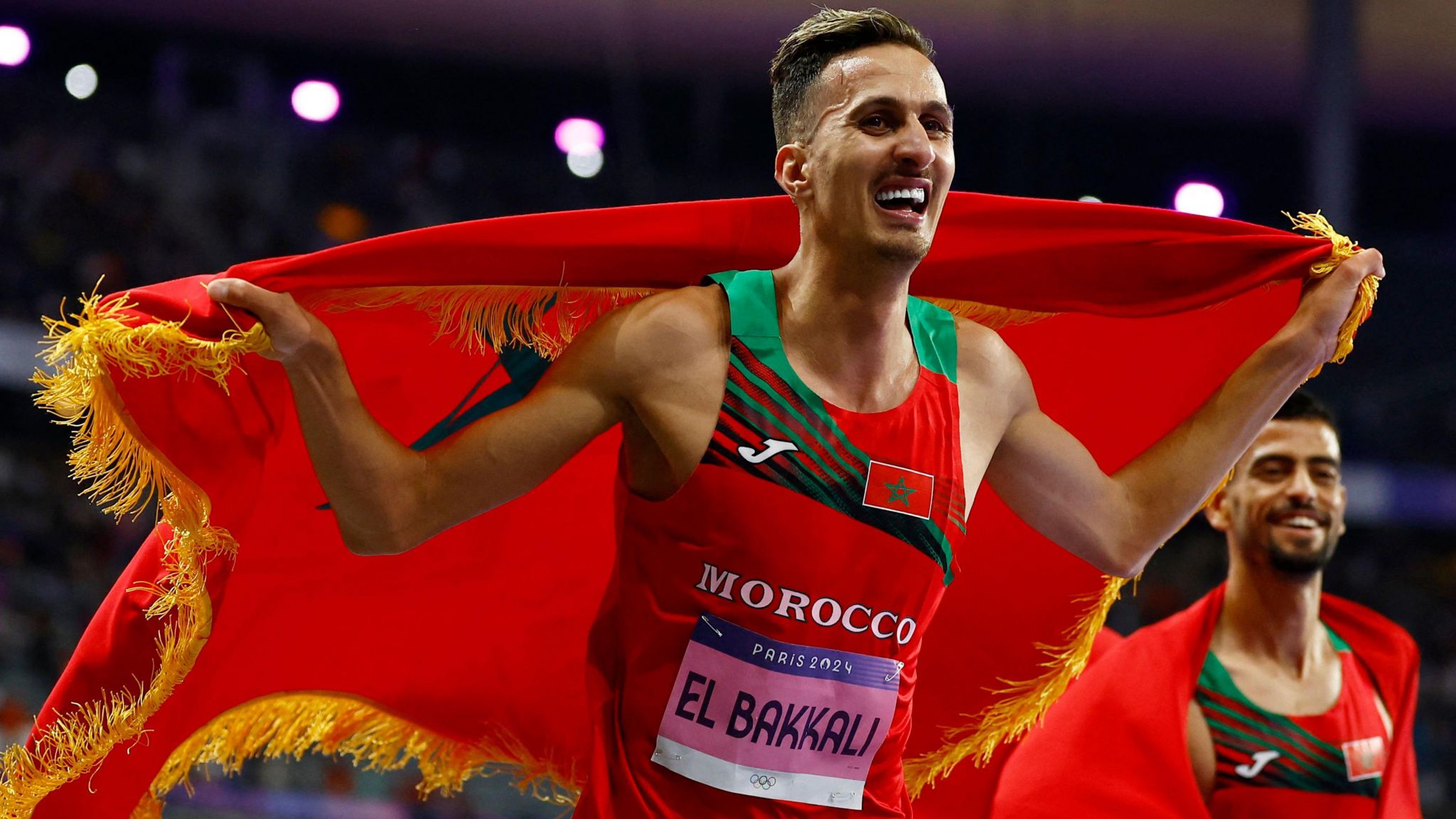 Soufiane El Bakkali smiles as he runs along celebrating his victory with a Moroccan flag with gold tassles trailing behind him