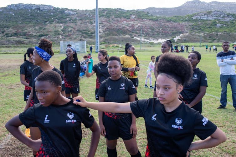 A group of girls in black sports uniforms are gathered on a sports field. In front, a girl with an Afro rests her hand on another girl's shoulders. There are mountains in the background.