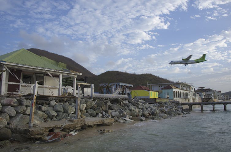 A large passenger plane approaches an island where houses built right to the water's edge are damaged from a recent hurricane.