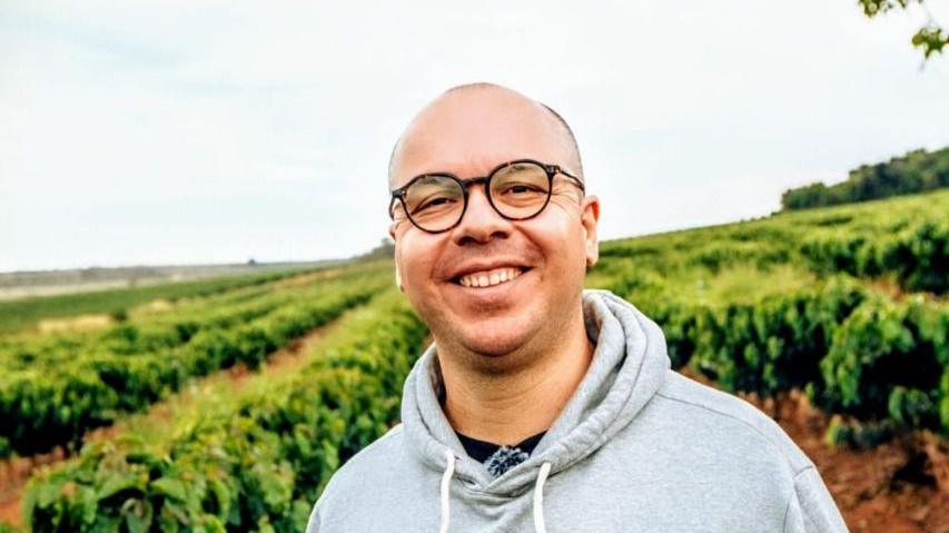Photo of coffee trader Hugo Portes, dressed in a light grey hoodie, standing in front of a coffee farm.

