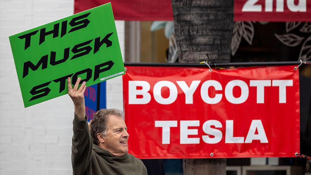A man holds a placard saying "This Musk Stop" in front of a banner reading "Boycott Tesla"