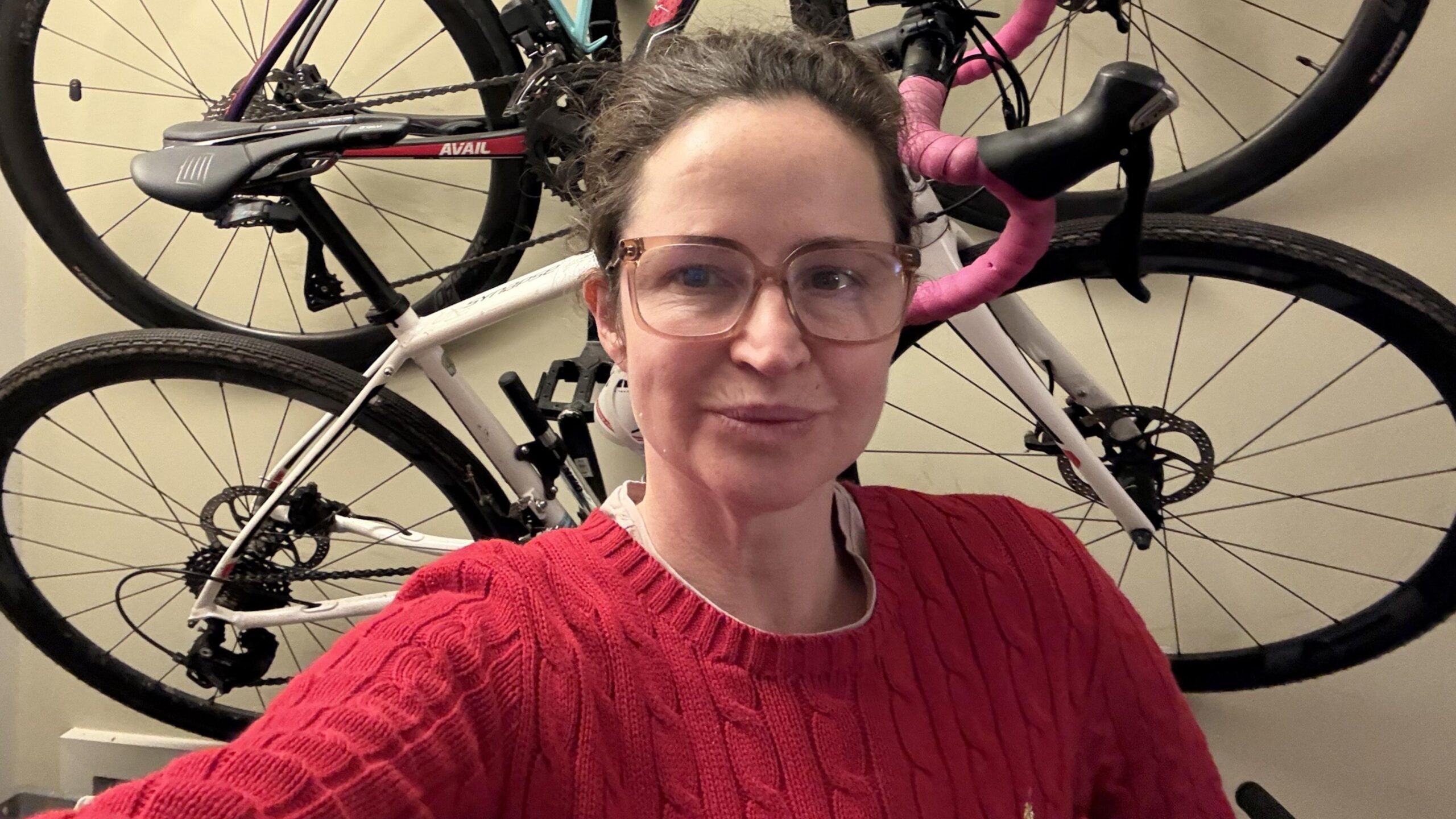 Wearing a red jumper and glasses, Kate Corden, stands in front of bicycles hanging on a wall.