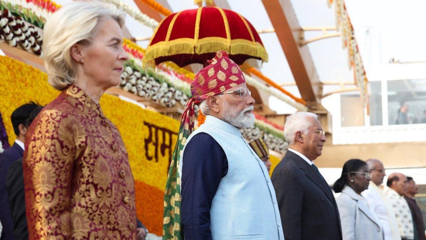 Leaders stand in a line on a decorated stage at an outdoor ceremony. From left to right are a European woman leader in a red patterned jacket, Indian Prime Minister Narendra Modi wearing a traditional turban and light blue vest, a European male leader in a dark suit, and Indian President Droupadi Murmu in a light-coloured coat, with floral decorations and officials visible in the background