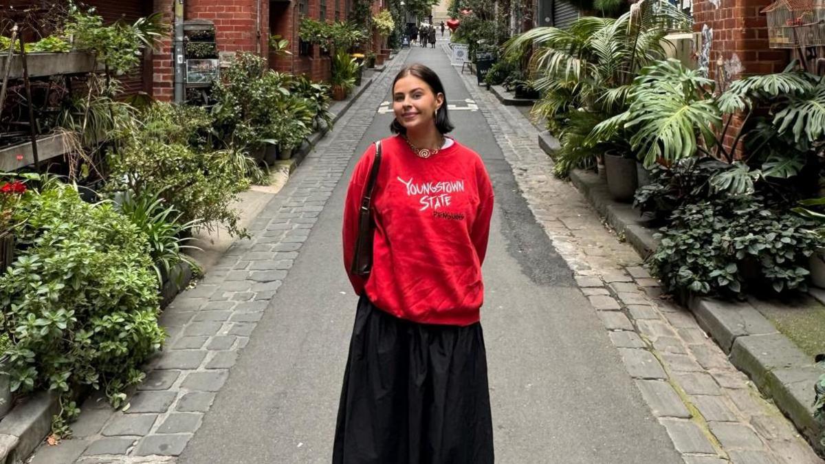 A young woman standing in a street. She is wearing a red jumper and a long black skirt. She has dark hair shoulder length hair and is standing in the middle of a street. It is narrow  with large green plants on either side of the small area of tarmac.