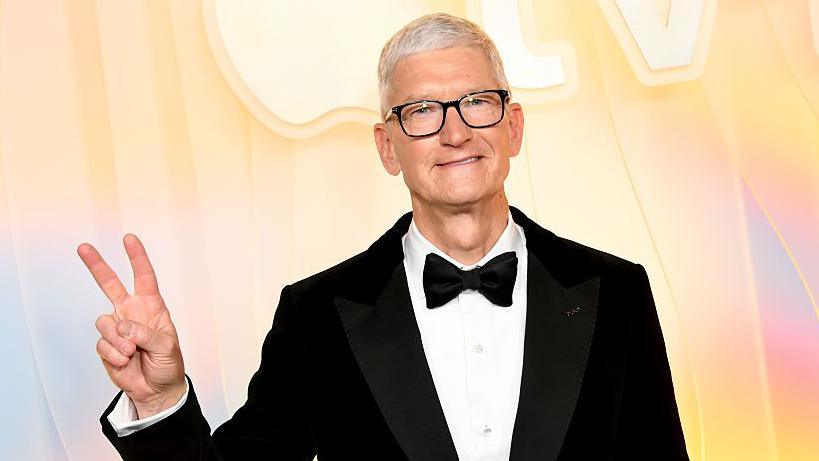Tim Cook wears a tuxedo and makes the "peace" sign as his stands in front of a yellow, blue and pink background reading Apple TV plus