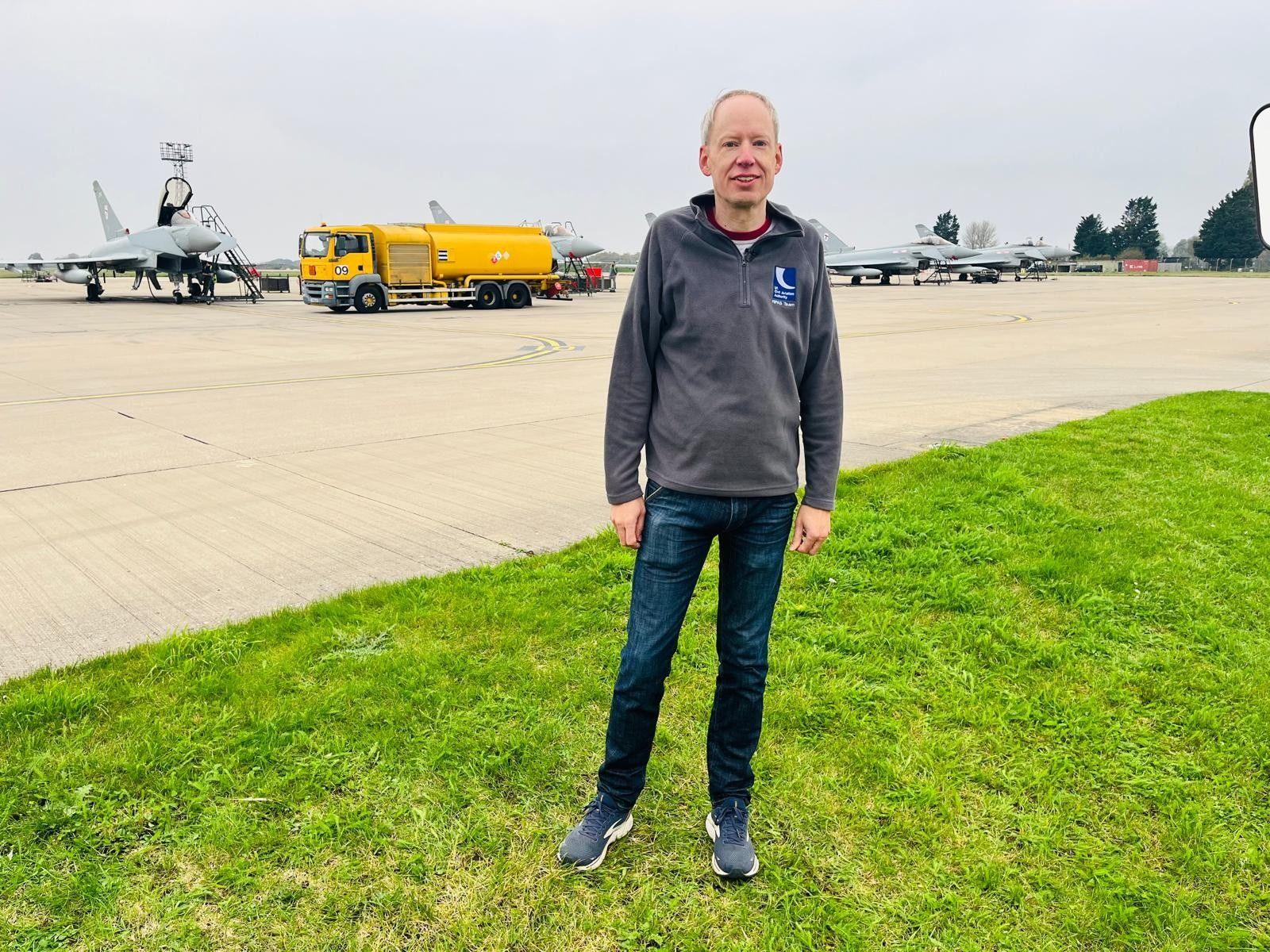 A man standing on grass in front of a fighter jet on an airfield.